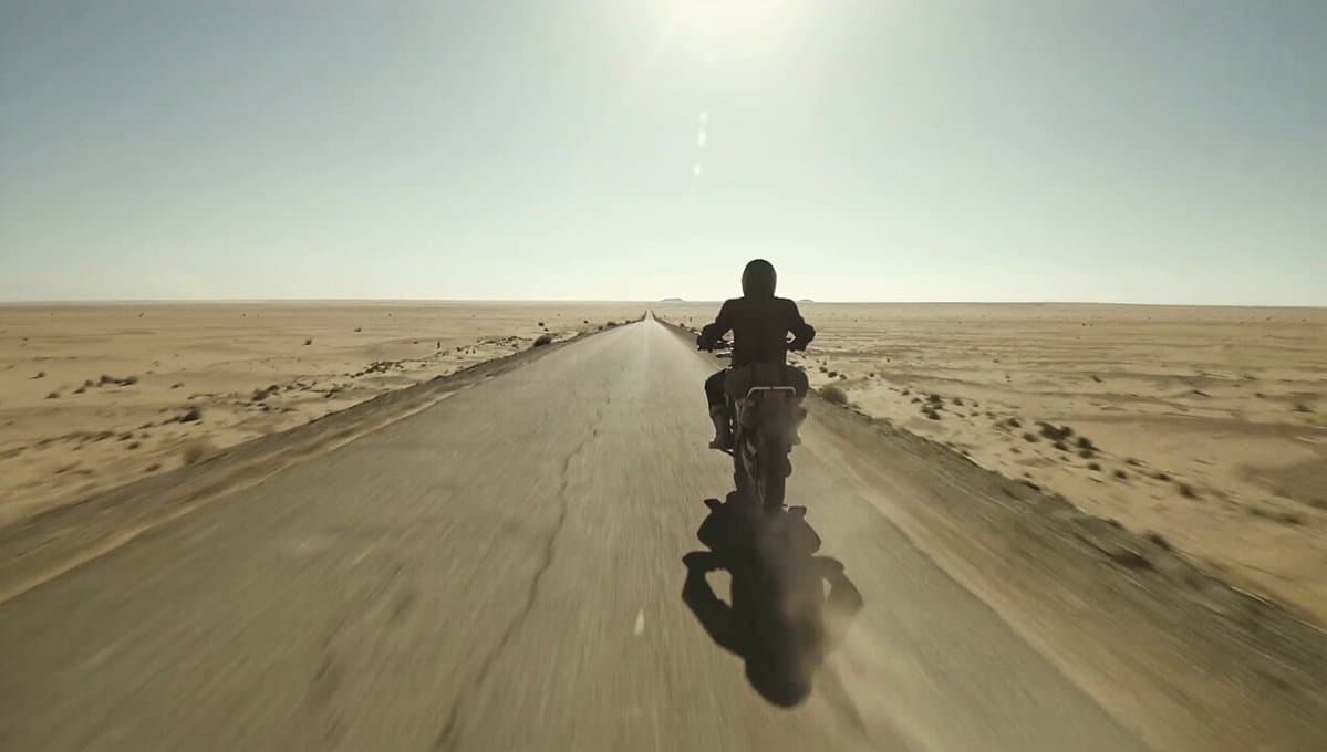 A motorbike rider crosses a remote desert road in brutal afternoon heat. The cam
